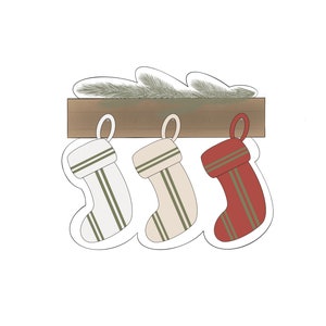 May include: Three Christmas stockings hanging from a wooden shelf with a sprig of greenery. The stockings are white with green stripes, beige with green stripes, and red with green stripes.