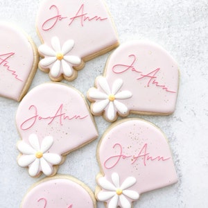 May include: Five pink heart-shaped cookies decorated with white daisies and the name "Jo Ann" written in pink cursive.
