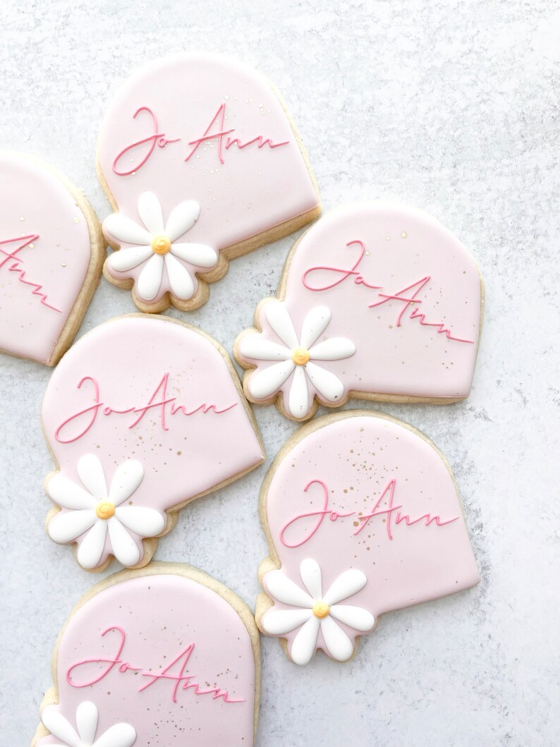 May include: Five pink heart-shaped cookies decorated with white daisies and the name "Jo Ann" written in pink cursive.