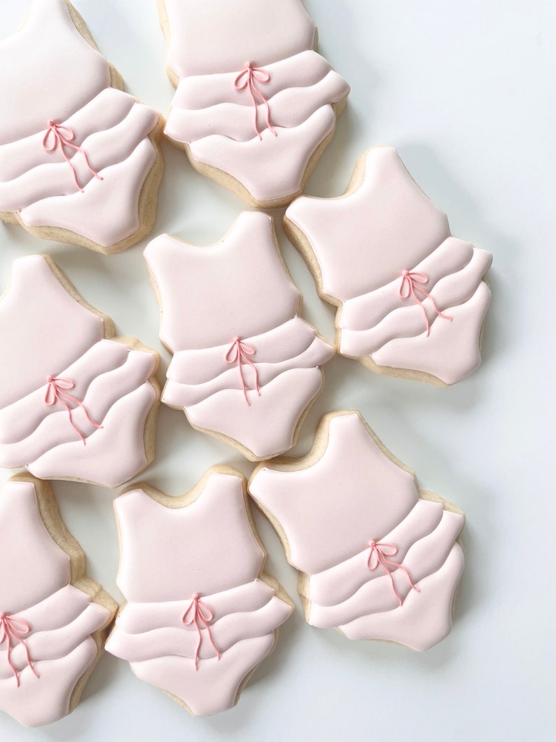 May include: Nine pink sugar cookies decorated with white icing and pink bows. The cookies are shaped like a one-piece swimsuit.