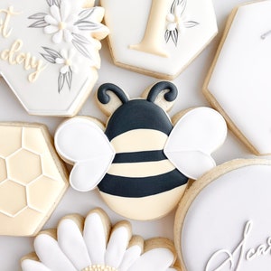 May include: A set of decorated sugar cookies featuring a bee, a daisy, a honeycomb, and the number one. The cookies are decorated with white, black, yellow, and gray icing.