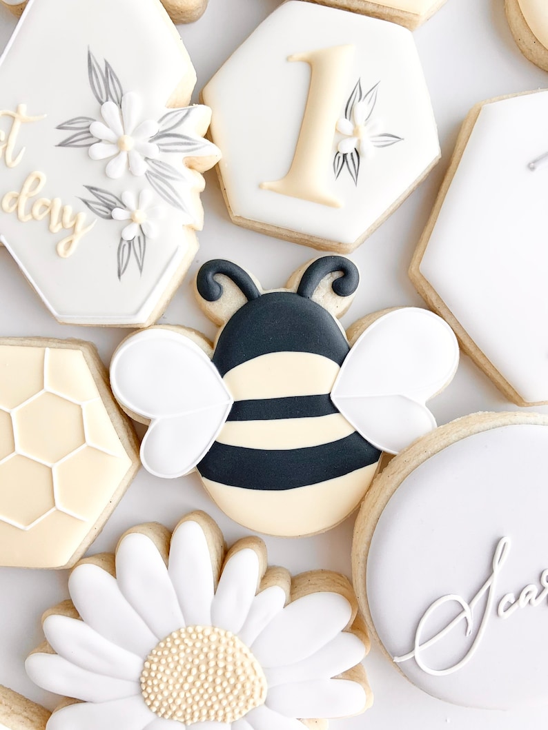 May include: A set of decorated sugar cookies featuring a bee, a daisy, a honeycomb, and the number one. The cookies are decorated with white, black, yellow, and gray icing.