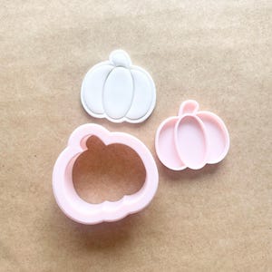 May include: Three pumpkin-shaped cookie cutters in white and pink. The cutters are made of plastic and are designed for baking. The shapes include a solid pumpkin, a pumpkin outline, and a smaller solid pumpkin.
