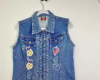 May include: A blue denim vest with a zipper closure. The vest has two smiley face patches with red, white, and blue hats and a heart-shaped patch with an American flag design. The vest also has the word "Good" written on it.