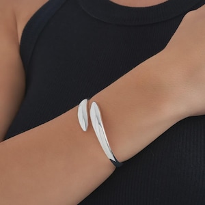 Może przedstawiać: Srebrna bransoletka typu cuff o zakrzywionym designie. Bransoletka posiada dwa końce w kształcie liści, które lekko na siebie zachodzą. Biżuteria jest noszona na nadgarstku, na tle czarnego topu.