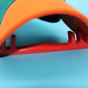 May include: A red plastic hat rack with three slots to hold baseball caps. The rack is designed to hold the brim of the hat.