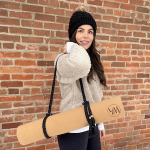 May include: A woman wearing a black beanie and a cream-coloured fuzzy sweater carries a cork yoga mat with a black strap and the letters "YM" printed on it.