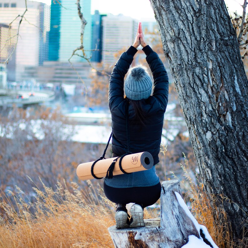 Pu&ograve; includere: Una persona con una giacca nera e un berretto grigio &egrave; seduta su un ceppo d'albero con le braccia alzate in una posa yoga. Tiene un tappetino da yoga marrone con un cinturino nero.