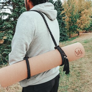 Pu&ograve; includere: Una persona che indossa una felpa con cappuccio grigia porta un tappetino da yoga marrone con un cinturino nero e un fiocco nero. Il tappetino ha un logo con le lettere "VM" su di esso.