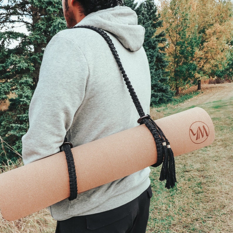 Pu&ograve; includere: Una persona che indossa una felpa con cappuccio grigia porta un tappetino da yoga marrone con un cinturino nero e un fiocco nero. Il tappetino ha un logo con le lettere "VM" su di esso.
