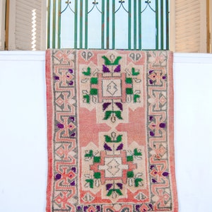 Puede incluir: Una alfombra rosa y crema con un patr&oacute;n geom&eacute;trico en verde, morado y gris. La alfombra est&aacute; colgada en una pared blanca.