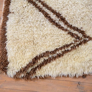 May include: A white shag rug with brown geometric designs. The rug has a long, shaggy pile and a fringe border.