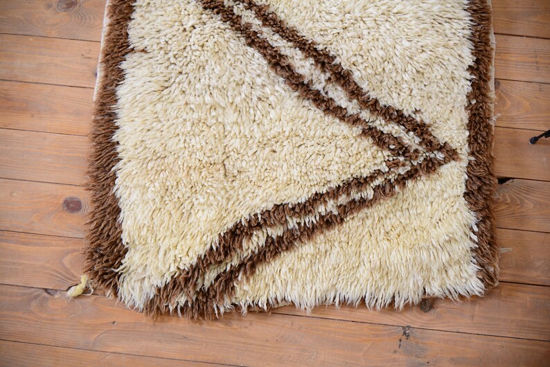 May include: A white shag rug with brown geometric designs. The rug has a long, shaggy pile and a fringe border.