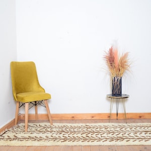 May include: A mustard yellow velvet chair with wooden legs sits on a white and brown patterned rug. A small black metal side table with a glass top sits in the background with dried pampas grass in a black vase.