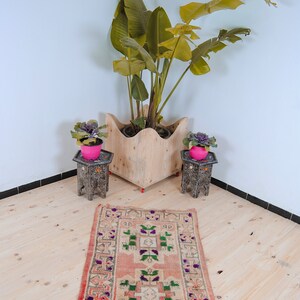 Puede incluir: Una alfombra rosa claro con un patr&oacute;n geom&eacute;trico en tonos de verde, morado y blanco. La alfombra est&aacute; colocada sobre un suelo de madera con dos mesas peque&ntilde;as negras y plantas en macetas.