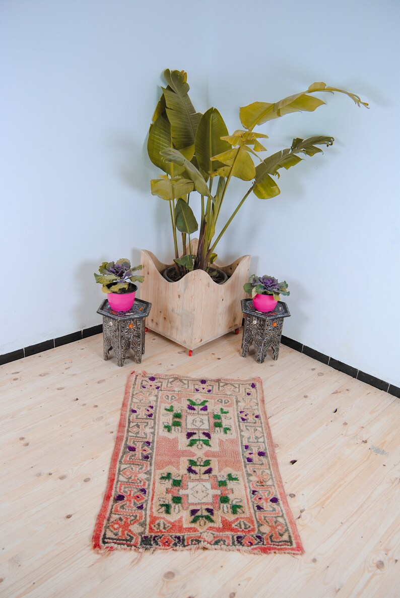 Puede incluir: Una alfombra rosa claro con un patr&oacute;n geom&eacute;trico en tonos de verde, morado y blanco. La alfombra est&aacute; colocada sobre un suelo de madera con dos mesas peque&ntilde;as negras y plantas en macetas.