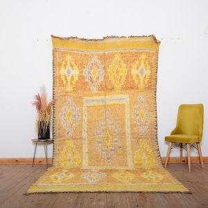 Puede incluir: Una alfombra rectangular grande con una combinación de colores amarillo y naranja. La alfombra presenta patrones geométricos en amarillo, gris y naranja. La alfombra se exhibe contra una pared blanca, con una silla amarilla y una mesa pequeña con un jarrón de flores secas.