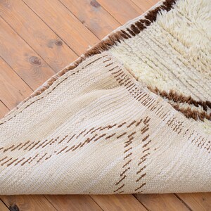 May include: A white and brown Moroccan rug with a shaggy texture. The rug has a geometric pattern with brown lines and a fringe border.