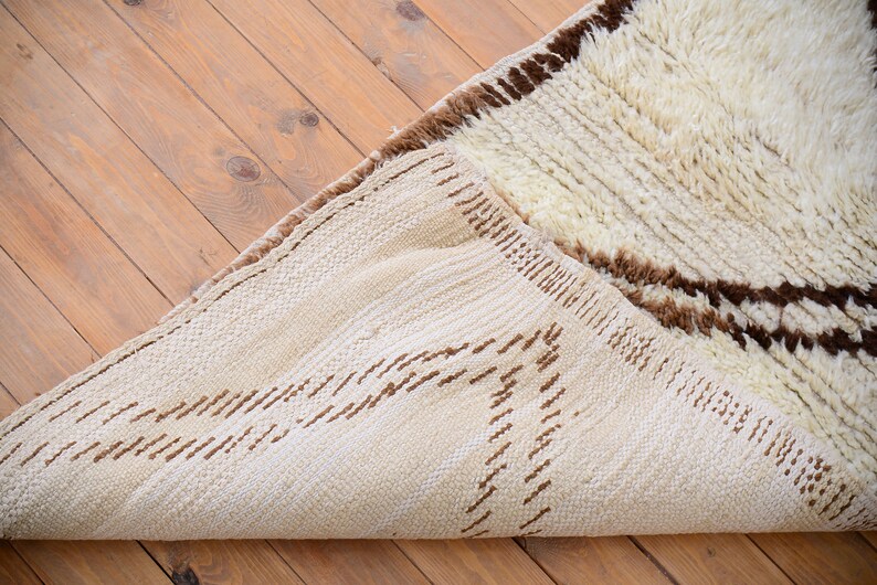 May include: A white and brown Moroccan rug with a shaggy texture. The rug has a geometric pattern with brown lines and a fringe border.