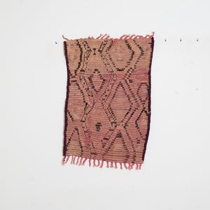 May include: A small, rectangular woven textile with a geometric diamond pattern in shades of pink, brown, and black. The edges are finished with a dark purple border and fringe. The textile is likely a decorative item.
