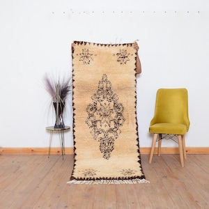 May include: A rectangular, beige rug with a central diamond-shaped pattern in dark brown. The rug has a fringed edge and is held up against a white wall. The rug's design features floral motifs.