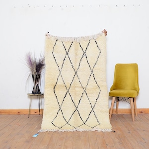 May include: Cream-colored, hand-woven rug with a geometric diamond pattern in black. The rug has a fringed edge and is displayed against a white wall. A yellow chair and a small table with a vase are in the background.