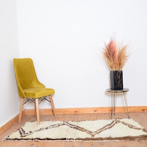 May include: A mustard yellow velvet chair with wooden legs sits in a corner of a room with white walls. A white and brown rug with a geometric pattern is on the floor. A small black vase with dried pampas grass sits on a gold metal side table.