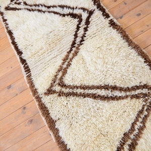 May include: A white shag rug with a brown geometric pattern. The rug has a long, rectangular shape and is perfect for adding a touch of texture and style to any room.