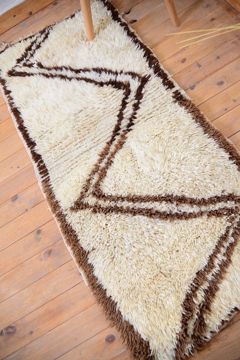 May include: A white shag rug with a brown geometric pattern. The rug has a long, rectangular shape and is perfect for adding a touch of texture and style to any room.