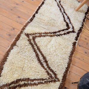 May include: A white shag rug with a brown geometric pattern. The rug has a long, rectangular shape and is made of soft, fluffy fibers.