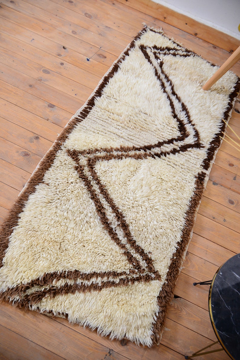 May include: A white shag rug with a brown geometric pattern. The rug has a long, rectangular shape and is made of soft, fluffy fibers.