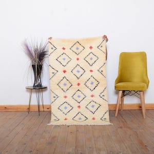 May include: Cream-colored rug with a diamond pattern. The diamonds are outlined in black and contain red, blue, and green accents. The rug is rectangular and has a fringed edge. The rug is being held up in front of a white wall.