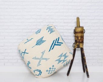 May include: A white floor cushion with blue geometric patterns. The cushion is sitting on a white tiled floor in front of a white brick wall. A decorative wooden stand with a gold metal top is in the background.