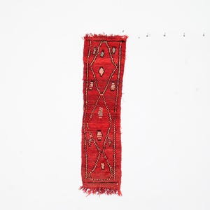 May include: A small, rectangular red rug with a geometric pattern. The rug features diamond and cross shapes in a lighter color, with a fringed edge. The rug is likely handmade and suitable for a small space.