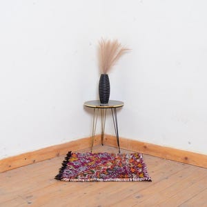 May include: A small, colorful rug with a fringed edge sits on a wooden floor. A black vase with dried flowers sits on a gold metal side table with three legs.