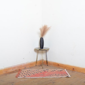 May include: A black vase with a textured surface and a bouquet of dried flowers sits on a small, round table with gold legs. A small, rectangular rug with a geometric pattern in black and coral is on the wooden floor.
