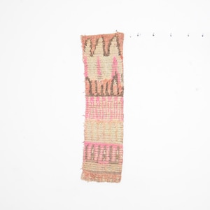 Puede incluir: Textil rectangular tejido con un patrón geométrico en tonos rosa, beige y marrón. El diseño presenta rayas verticales y formas repetidas. El textil tiene un borde con flecos y un aspecto texturizado.