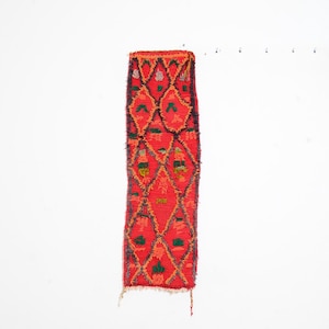 May include: A rectangular, woven textile with a vibrant red background and a repeating diamond pattern. The diamonds are outlined in orange and filled with green and yellow accents. The edges have a fringed finish.