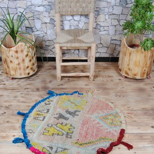 May include: A round, colorful rug with fringe, featuring a geometric pattern in shades of pink, blue, yellow, and white. The rug is placed on a wooden floor in front of a wooden chair and two potted plants.