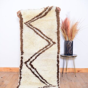 May include: A white and brown Moroccan rug with a geometric pattern. The rug is made of wool and has a soft, fluffy texture.