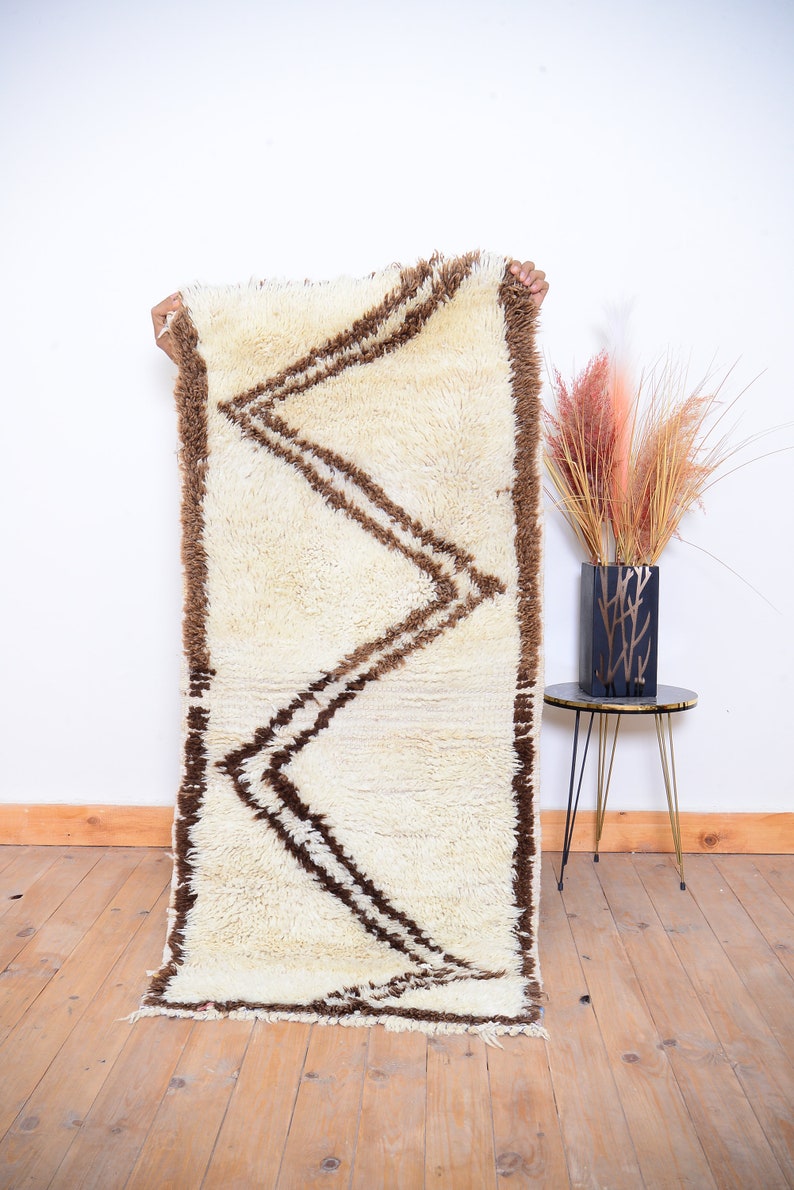 May include: A white and brown Moroccan rug with a geometric pattern. The rug is made of wool and has a soft, fluffy texture.