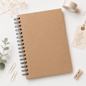 Può includere: Un taccuino a spirale con copertina in carta kraft marrone tinta unita, accompagnato da un rotolo di nastro adesivo con motivo a griglia, un'etichetta rettangolare con un disegno floreale nero, due graffette dorate e una piccola molletta in legno.