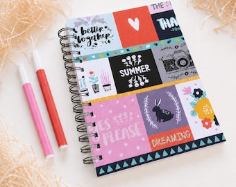 May include: A spiral-bound notebook featuring a multi-panel cover with various illustrations and text including "better together," "SUMMER," "YES PLEASE," and "DREAMING," accompanied by a pink marker and a red marker on the side.