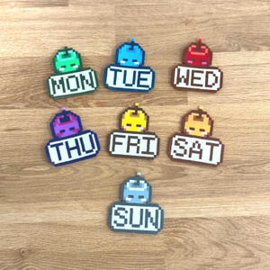 May include: A set of seven pixel art style day-of-the-week labels made from fused beads. Each label features a unique color, a small character icon at the top, and the text MON, TUE, WED, THU, FRI, SAT, or SUN in a bold, white, blocky font.