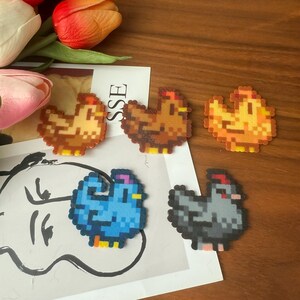 May include: A collection of pixelated chicken-shaped crafts in shades of brown, orange, blue, and grey. The crafts are displayed on white paper, with a floral arrangement in the background. Each chicken has yellow feet and a red comb.