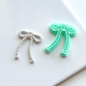 May include: Two plastic cookie cutters in the shape of a bow. The larger cutter is light green and the smaller cutter is white.