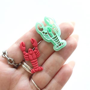 Puede incluir: Dos cortadores de galletas con forma de langosta, uno rojo y otro verde, se sostienen en una mano.