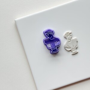 May include: Two cookie cutters in the shape of a woman's bust. One is purple and the other is white.