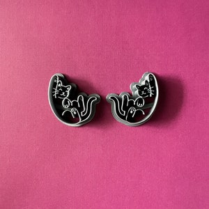 May include: Two grey metal cookie cutters shaped like cats curled up in a sleeping position. The cats have black eyes and noses.