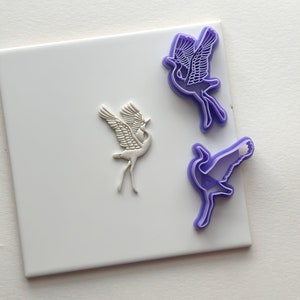 May include: Two purple plastic cookie cutters in the shape of a crane, one with its wings spread and the other with its wings folded. The cutters are on a white tile surface.